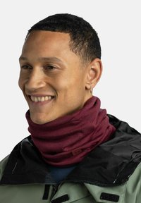 Burgundy neck gaiter made of soft, textured fabric, worn over a jacket with a black collar. Features a small brand logo tag.