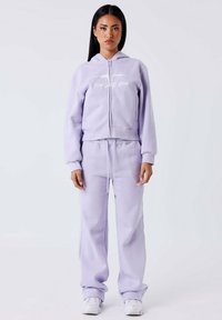 Felpa con zip lavanda, dotata di tasche frontali e pantaloni coordinati, con vita regolabile tramite coulisse e dettagli del logo discreti. Indossata con sneakers.