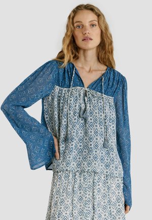 Frau mit welligem blondem Haar trägt eine blaue, gemusterte Bluse mit langen Ärmeln, die unter einem weißen und blauen, gemusterten ärmellosen Oberteil und Rock getragen wird.