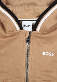 Haut à zip beige avec une bordure rayée noire et blanche, comportant une fermeture éclair à l'avant et un logo "BOSS" blanc sur la poitrine gauche. Texture de tissu douce.