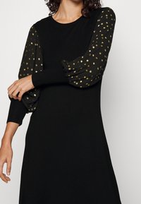 Svart klänning med långa puffy ärmar gjorda av genomskinligt tyg med metalliska guldprickar. Helt svart kropp med rundad halsringning.