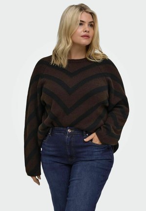 Mujer rubia con cabello de longitud media que lleva un suéter con patrón de chevron marrón y negro y vaqueros azul oscuro con una mano en el bolsillo.