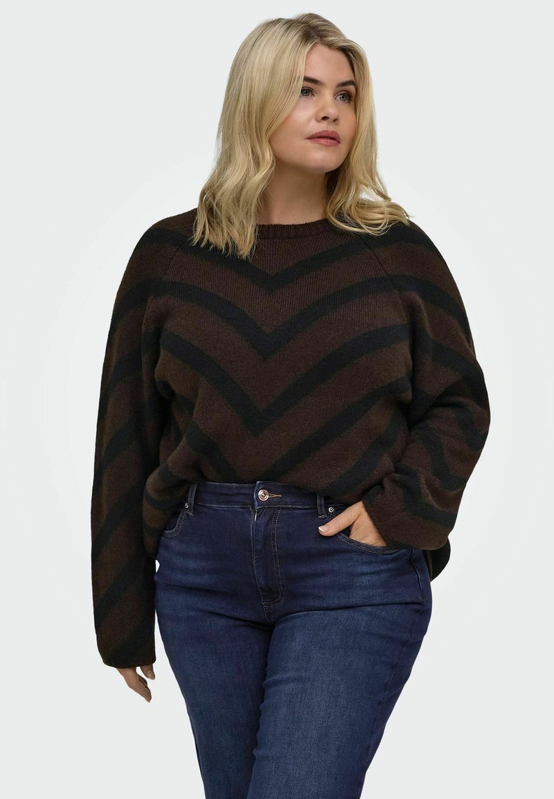 Femme blonde aux cheveux mi-longs portant un pull chevron marron et noir et un jean bleu foncé avec une main dans la poche.