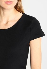 Svart kortärmad topp med rundad halsringning. Slätt, stretchigt material med en figurnära design som visar axlarna och övre delen av bröstet.