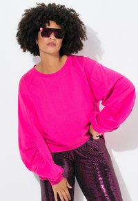 Sudadera rosa con mangas abullonadas combinada con pantalones rosas brillantes y con lentejuelas. La modelo lleva gafas de sol reflectantes. El fondo es blanco liso.