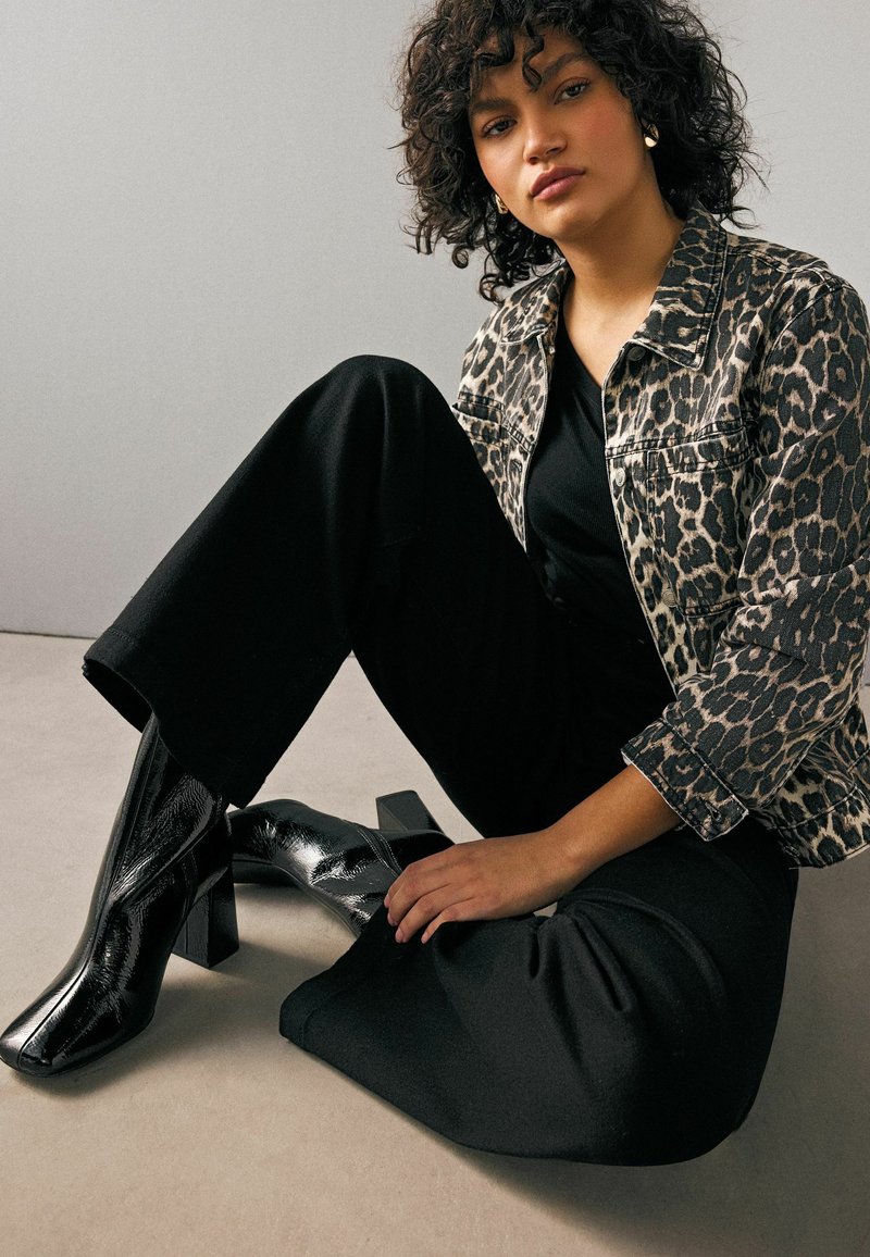 Giacca di jeans con stampa leopardata sopra una maglietta nera, abbinata a pantaloni neri a zampa e stivaletti neri lucidi con tacco. Capelli ricci, mani che riposano sulla gamba.
