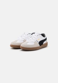 Fehér és bézs sneakers, amelyek velúrból és bőrből készültek, fekete díszítéssel, kerek orral és barna, mintás gumitalppal.