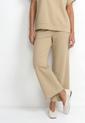 Person iført beige bukser med vide ben med matchende top og hvide perforerede slip-on sko mod en hvid baggrund.