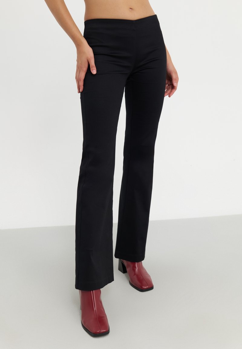 Czarne spodnie dzwony z elastycznego materiału, o gładkiej teksturze i wysokim stanie. Na nogach czerwone botki do kostki na grubej szpilce.