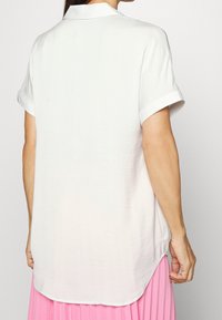 Camicia bianca a maniche corte con colletto, caratterizzata da una vestibilità comoda e un orlo curvo. Il tessuto appare liscio e leggero.