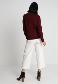 Maglione in burgundy lavorato a maglia con vestibilità ampia, abbinato a pantaloni bianchi a gamba larga e stivali neri alla caviglia con dettagli in legno.