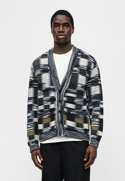 Άνδρας που φορά ένα πουκάμισο τύπου καρντιγκάν με κουμπιά σε πρότυπο μαύρο, άσπρο, ναυτικό και ελιά, πάνω από ένα λευκό πουκάμισο και μαύρο παντελόνι, στέκεται μπροστά σε ένα απλό φόντο.
