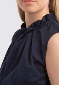 Donkerblauwe mouwloze blouse met een rimpelige hoge hals, zachte textuur, subtiele plooien aan de voorkant en een verborgen rits.