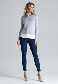 Grijze langemouwen top met een witte zoom, gecombineerd met donkerblauwe skinny jeans en lichtgrijze hoge hakken, tegen een neutrale achtergrond.