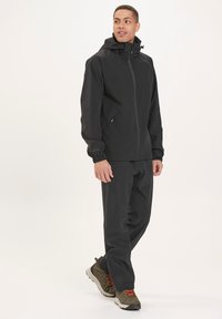 Sort vandtæt jakke med hætte, frontlynlås og justerbare manchetter. Bæres med matchende bukser og grå-grønne sko med orange snørebånd.