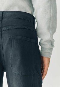 Dark denim pants feature a back pocket, visible stitching details, and a high waist. A light grey sweater is partially visible.