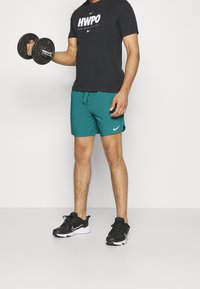 Man som bär en svart "HWPO" T-shirt, teal Nike-shorts och svarta Nike-sneakers, hållande en hantel i sin högra hand.