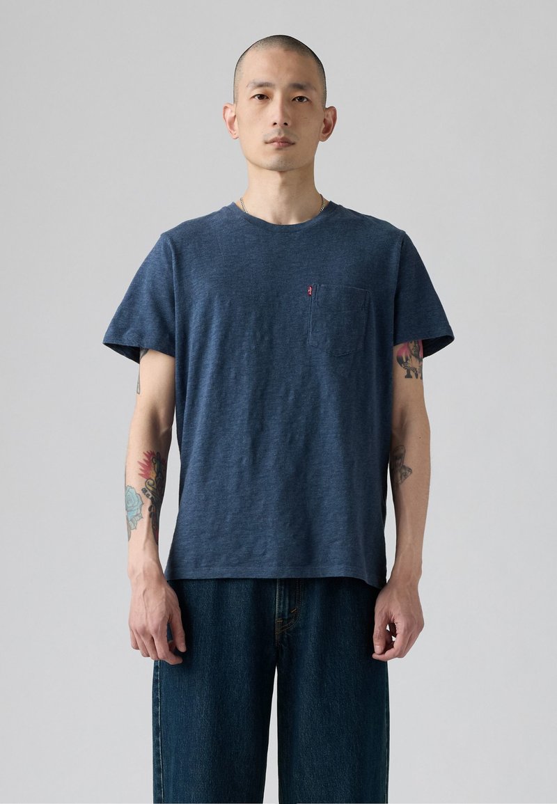 Mann mit Tätowierungen, der ein marineblaues T-Shirt mit kurzen Ärmeln und dunkle Jeans vor einem schlichten hellgrauen Hintergrund trägt.