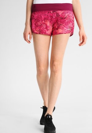 Frau trägt rosa gemusterte Sportshorts mit breitem bordeauxrotem Bund und schwarzen Laufschuhen vor einfarbigem Hintergrund.