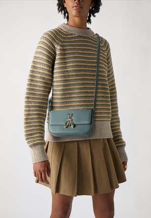 Maglione a righe in beige e giallo, abbinato a una gonna plissettata color khaki. Una borsa a tracolla in pelle verde presenta una chiusura decorativa a forma di insetto.