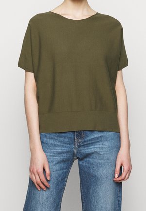 Personne portant un haut olive à manches courtes larges et un jean bleu en denim, debout devant un fond clair uni.