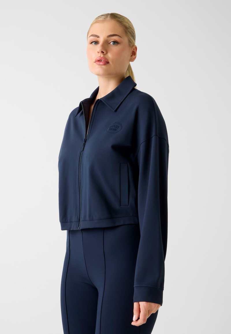 Giacca corta blu navy in tessuto liscio con zip frontale e colletto. Presenta un logo sul petto sinistro e tasche laterali.