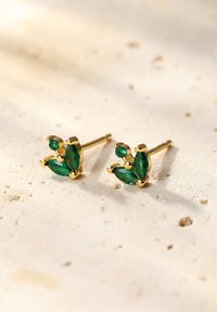 Pendientes de oro con piedras preciosas verdes en forma de hoja y piedras de acento más pequeñas, colocadas sobre una superficie texturizada.