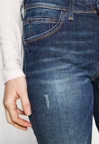 Tmavě modré džíny z denimu s patrnými vybledlými místy a mírným opotřebením. Šité detaily v oranžové barvě. Ruka modelky spočívá na bocích.
