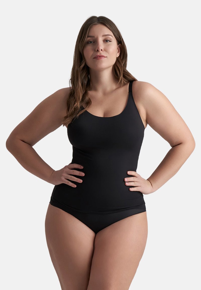 Vrouw met lang haar in een zwarte tanktop en zwarte onderbroek, staand met de handen op de heupen tegen een effen achtergrond.