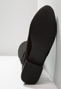 Schwarzer Schuh mit niedrigem Blockabsatz, der mit der Sohle nach oben an einer weißen Oberfläche und Wand lehnt, zeigt die strukturierte Sohle und den glatten Absatzboden.