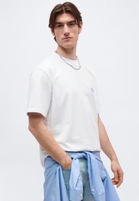 Νέος άνδρας που φοράει λευκό T-shirt και ανοιχτό μπλε τζιν, με ένα μπλε ριγέ πουκάμισο δεμένο γύρω από τη μέση του, με τα χέρια χαλαρά.