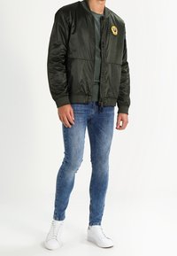 Oliivinvihreä pommittaja-takki, jossa on vetoketjukinna ja ribatty hihansuut, varustettuna logopatchilla. Yhdistetty vaaleansinihin farkkuihin ja valkoisiin lenkkareihin.