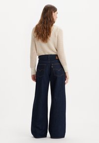 Vida mörka jeans i denim med hög midja och två bakfickor, kombinerade med en krämfärgad stickad tröja med öppet mönster.