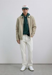 Jachetă bej ușoară peste un hoodie verde închis, blugi crem și adidași albi. Căciulă de baseball cu detalii în roșu și verde.