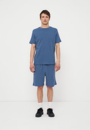 Moški stoji na belem ozadju, oblečen v modro majico s kratkimi rokavi in ustrezne kratke hlače, bele nogavice in črne superge.