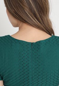 Tyrkysové krajkové šaty s texturovaným vzorem, krátkými rukávy a zadním zipem. Hrbáčkový výstřih a látka obsahuje složité detaily.