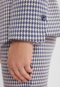 Rękaw marynarki w wzór houndstooth w kolorze granatowym i białym, z pojedynczym granatowym guzikiem, wykonany z teksturowanej tkaniny z widocznym przeszyciem.