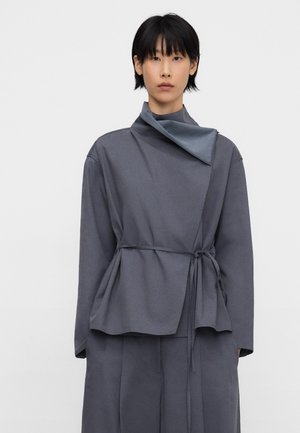 Person trägt ein locker sitzendes, langärmliges graues Oberteil mit hohem Kragen und verstellbarem Bindeband an der Taille, kombiniert mit passenden grauen Hosen.