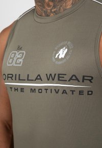 Olijfgroene mouwloze sportschoolshirt van rekbare stof, met witte grafische tekst en een cirkelvormig logo aan de voorkant.