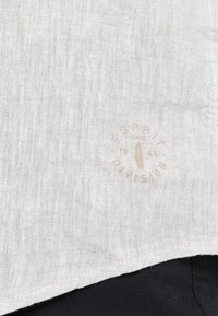 Tela de lino blanca con un acabado texturizado. Presenta un logotipo circular en marrón claro que dice "ESPRIT DIVISION 1968" cerca del borde inferior.