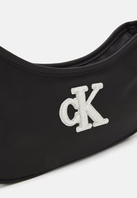 Čierna látková taška s elegantným dizajnom, obsahujúca bielo vyšívané logo "cK". Mäkká textúra a zakrivený tvar pre jednoduché prenášanie.