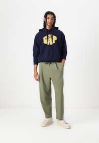 Hanorac bleumarin cu logo-ul galben "GAP"; pantaloni relaxați de un verde măsliniu; adidași crem; ținută casual cu un design minimalist.