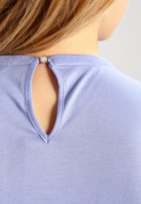 Vaaleanpunainen t-paita, jossa on selässä avainreikäkuvio, pieni hopeinen nappidetalji kaula-aukossa ja sileä kangaspinta.