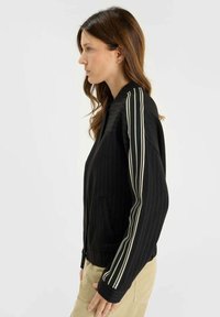 Kvinna i beige byxor som bär en svart zip-up jacka med vita vertikala ränder på ärmarna, stående i profil mot en enfärgad bakgrund.