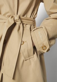 Trench-coat beige noué à la taille avec une ceinture, montrant un poignet boutonné avec le nom de la marque brodé sur la manche.