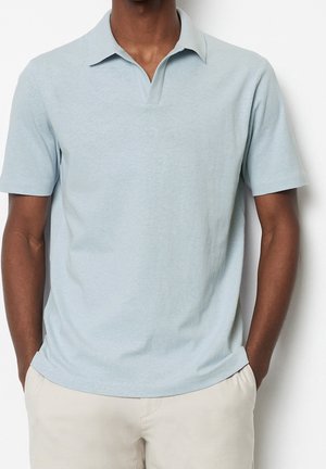 Homme portant un polo bleu clair à manches courtes et un pantalon beige, les mains dans les poches, sur un fond blanc uni.