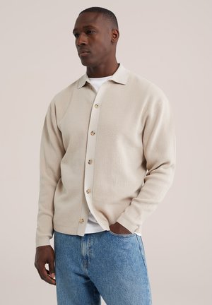 Homme portant un cardigan beige boutonné sur une chemise blanche et un jean bleu, debout devant un fond clair uni avec une main dans la poche.