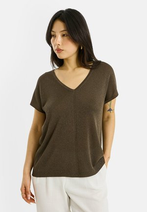 Femme aux cheveux noirs mi-longs portant un haut marron à manches courtes et col en V, ainsi qu'un pantalon blanc, regardant vers la gauche avec des tatouages visibles sur son bras.