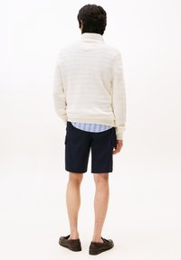 Helle Strickjacke mit gerippter Textur, über einem blau gestreiften Hemd getragen, kombiniert mit dunklen navy Cargo-Shorts und dunklen Schuhen.