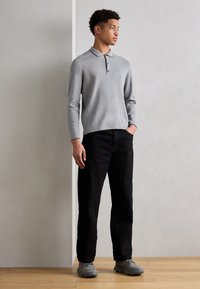 Polo shirt gris à manches longues avec un col et trois boutons, associé à un pantalon noir et des baskets grises, se tenant sur un sol en bois.